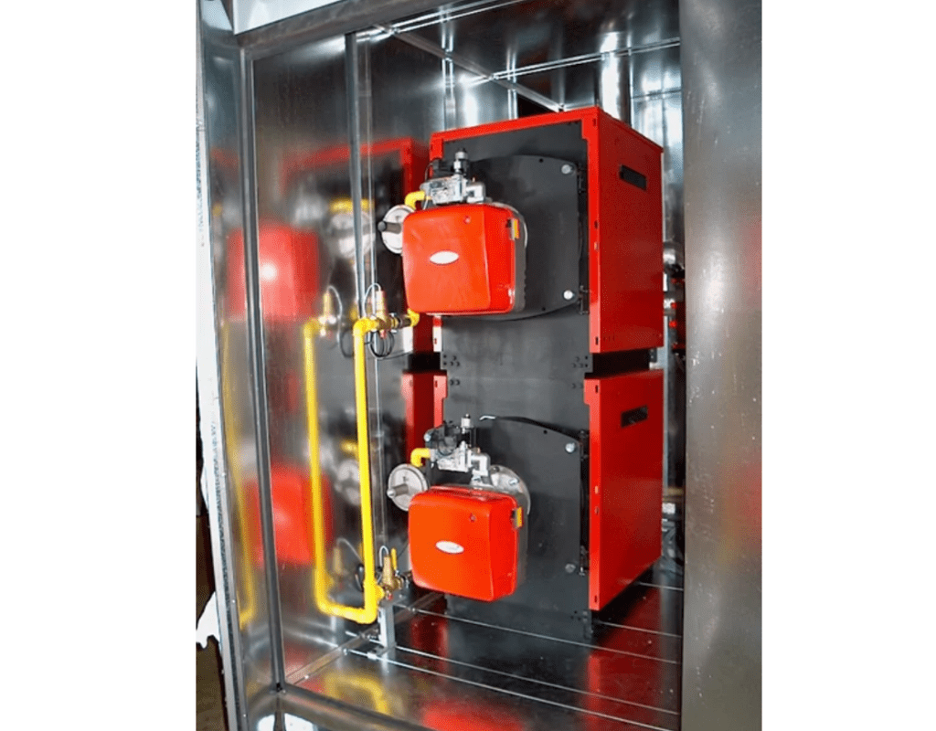 Deux brûleurs industriels rouges montés verticalement à l'intérieur d'une enceinte métallique, avec des tuyaux jaunes et des vannes. Ils font partie d'une chaudière eau chaude en container extérieur
