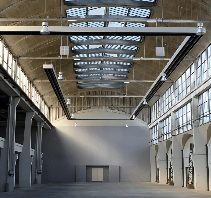 Grands radiants de chauffage fixe suspendus au plafond d'un vaste espace industriel éclairé par des puits de lumière.