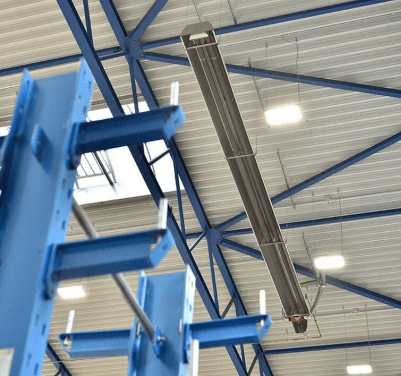 Radiant de chauffage fixe (tube radiant gaz), long et tubulaire, suspendu au plafond d'un espace industriel avec une structure métallique bleue.